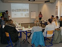 Tischkreis der Lehramtsanwärter Man sieht mehrere Personen, welche an Tischen in einer U-Formation sitzen. Zwei Personen stehen und stellen Präsentationsfolien vor. Auf einem Flipchart steht herzlich willkommen.