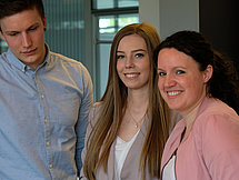 Zwei Frauen und ein Mann stehen an einem Tisch. Die Frauen schauen in die Kamera, während der Mann ein auf dem Tisch liegendes Heft liest.