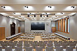 The picture shows a big conference room featuring a big screen, light grey materials and wooden details.