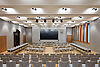 L'immagine mostra una grande sala conferenze con un grande schermo, materiali grigio chiaro e dettagli in legno.