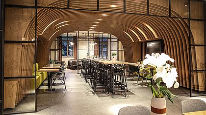 Hotelrestaurant mit hohen und niedrigen Tischen unter einer offenen, bogenförmigen Deckenkonstruktion aus Holzträgern