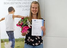 Abschlussfoto Jessica Richter Eine junge Dame präsentiert stolz ihr Prüfungszeugnis und einen Blumenstrauß.