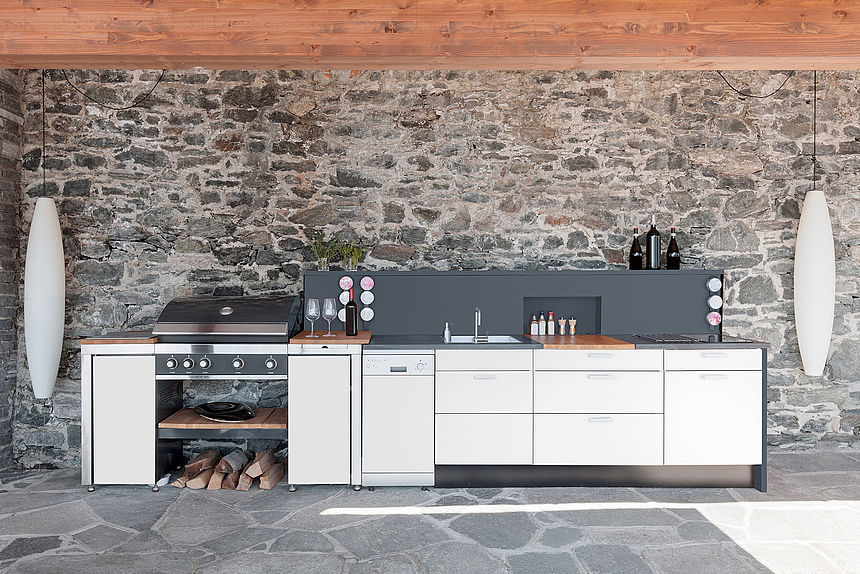 XTerior compact from Pfleiderer in the outdoor kitchen An outdoor kitchen made of XTerior compact in white with a grey back wall in front of a stone wall.