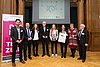 Acht Personen posieren für ein Gruppenbild in einer Halle mit hölzernem Boden und hölzernen Wänden. Links ist ein Banner der "Schulewirtschaft" zu sehen.