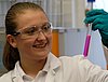 A young woman in a lab coat carrying and looking at a purple substance in a glass tube