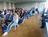 Man sieht etwa 30 Personen von vorne, welch in Stuhlreihen sitzen und einer Person zuhören, welche nicht im Bild zu sehen ist.