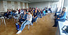 Man sieht etwa 30 Personen von vorne, welch in Stuhlreihen sitzen und einer Person zuhören, welche nicht im Bild zu sehen ist.