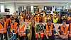 Etwa 20 Auszubildende in orangenen Westen posieren für ein Gruppenbild in einer Werkstatt