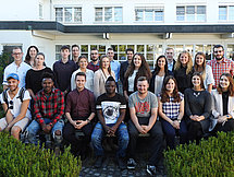 Gruppenfoto Man sieht etwa 25 junge Leute welche auf bzw. hinter einer Bank für ein Gruppenfoto posieren. Im Hintergrund ist ein Gebäude zu sehen.