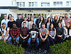 Man sieht etwa 25 junge Leute welche auf bzw. hinter einer Bank für ein Gruppenfoto posieren. Im Hintergrund ist ein Gebäude zu sehen.