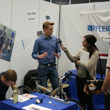 Interview auf Messestand Eine Frau interviewt einen jungen Mann mit einem Mikrofon auf einem Messestand