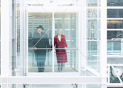 Ein Rentnerpaar fährt glücklich lächelnd in einem gläsernen Aufzug.