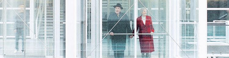 Ein Rentnerpaar fährt glücklich lächelnd in einem gläsernen Aufzug.