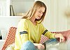 The picture shows a young, blonde woman sitting in a chair cross-legged. She's reading a magazine. The background shows a book shelf with decor.