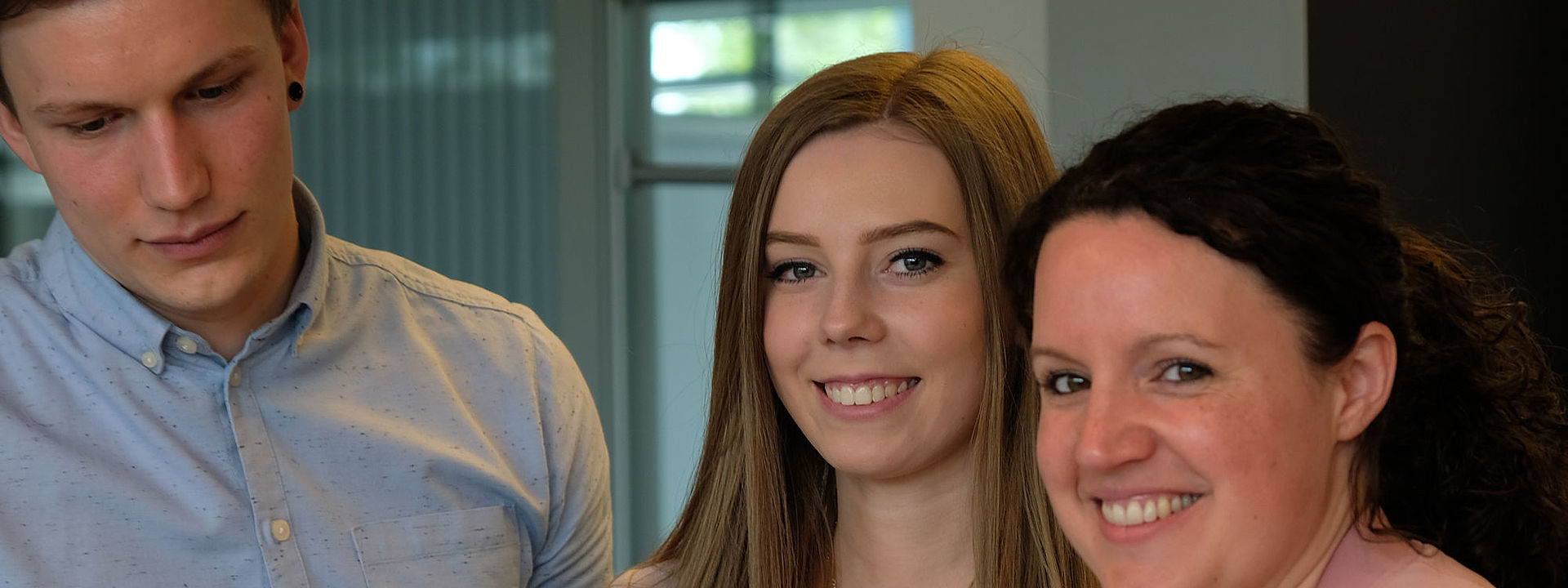 Symbolbild Industriekaufleuter Das Bild zeigt 2 Frauen und 1 Mann, die an einem Tisch stehen. Die Frauen schauen direkt in die Kamera, während der Mann ein Buch liest, das auf dem Tisch liegt