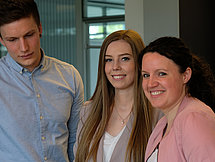 Symbolbild Industriekaufleuter Das Bild zeigt 2 Frauen und 1 Mann, die an einem Tisch stehen. Die Frauen schauen direkt in die Kamera, während der Mann ein Buch liest, das auf dem Tisch liegt