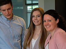 Representative image Salespeople The picture shows two women and a man standing at a table. The two women smile into the camera while the man is reading a booklet that's laying on the table