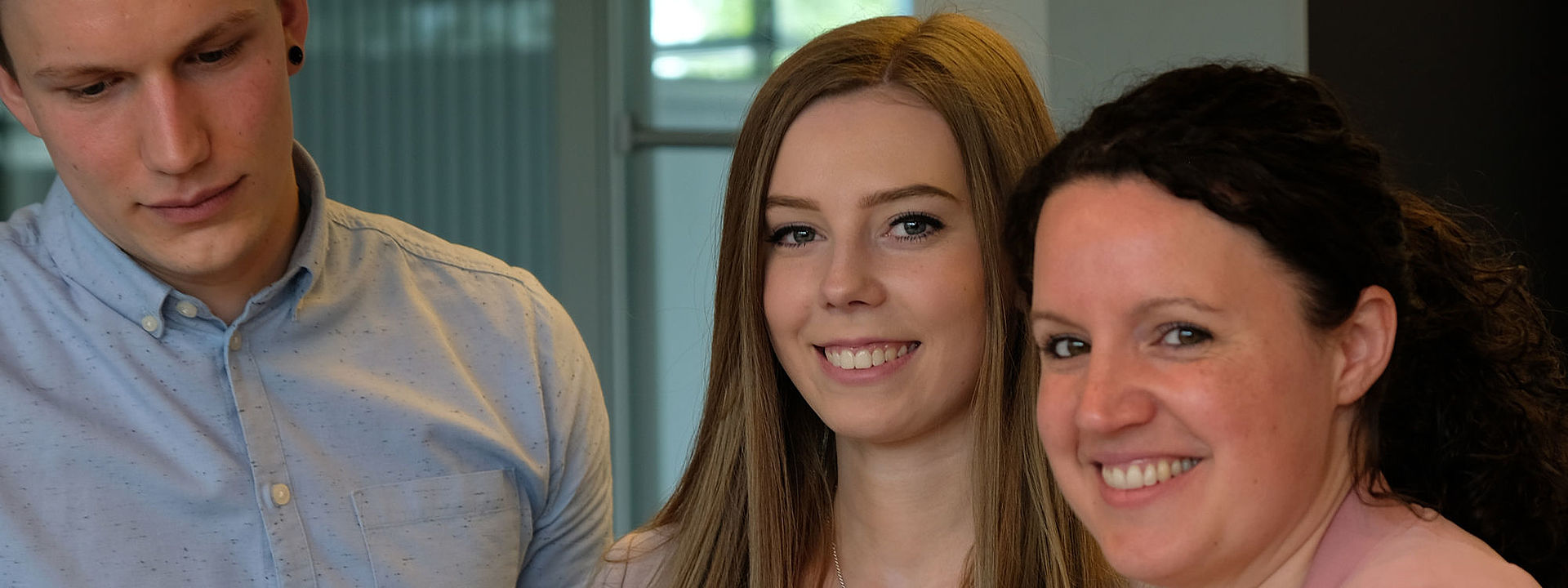 Symbolbild Industriekaufleute Das Bild zeigt zwei Frauen und einen Mann, die an einem Tisch stehen. Die beiden Frauen lächeln in die Kamera, während der Mann eine Broschüre liest, die auf dem Tisch liegt