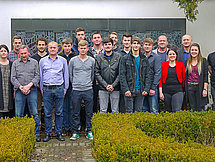 Gruppenfoto Etwa 20 Personen posieren draußen für ein Gruppenfoto vor einer Wand