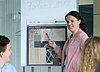 The picture shows a young lady in front of a flipchart, on which she explains something to the graduates present.