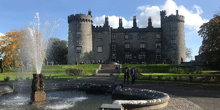 Schloss in Dublin Man sieht einen alten Brunnen vor einem imposanten britischen Schloss an einem sonnigen Tag.