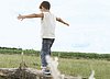 The picture shows a small boy in jeans and a white shirt from behind, balancing in nature on a meadow on a tree trunk. Above him the blue sky, in the background you can see the vastness of nature.