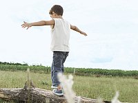Social sustainability The picture shows a small boy in jeans and a white shirt from behind, balancing in nature on a meadow on a tree trunk. Above him the blue sky, in the background you can see the vastness of nature.