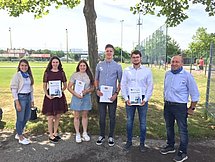 @Pfleiderer Neumarkt GmbH 4 Auszubildende zeigen stolz ihre Prüfungsurkunden. Neben ihnen stehen ihre Ausbilder. Sie befinden sich an einem Sportplatz.