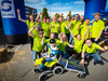 Etwa 15 Erwachsene in einheitlicher, neon-grüner Kleidung und ein Junge auf einem Bobbycar mit Solarzellen posieren für ein Gruppenbild. Sie jubeln.