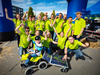Etwa 15 Erwachsene in einheitlicher, neon-grüner Kleidung und ein Junge auf einem Bobbycar mit Solarzellen posieren für ein Gruppenbild. Sie jubeln.