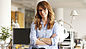 The picture shows a young lady with long red hair and glasses standing in an office in front of her desk smiling directly into the camera.