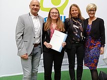Ehrung der Besten Vier Personen posieren auf einem grünen Teppich vor einem Banner der IHK Arnsberg. Sie tragen chique Kleidung.