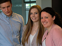 Zwei Frauen und ein Mann stehen an einem Tisch. Die Frauen lächeln in die Kamera, während der Mann ein Buch liest, das auf dem Tisch liegt.