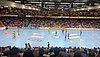 Man sieht eine Sporthalle mit voll besetzter Tribüne. Das Handballspiel scheint gerade vor dem Anpfiff zu stehen.