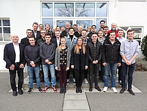 Gruppenbild der Auszubildenden und Ausbilder Man sieht eine Gruppe von Auszubildenden und Ausbildern, welche vor dem Eingang eines Gebäudes für ein Gruppenbild in Reihen stehen.