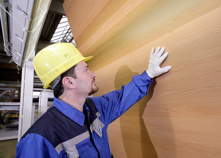 Representative image "Skilled Workers" A man wearing a yellow hard hat and blue protective clothing checks the coating on an upright light-colored wooden panel.
