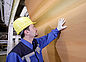 A man wearing a yellow hard hat and blue protective clothing checks the coating on an upright light-colored wooden panel.
