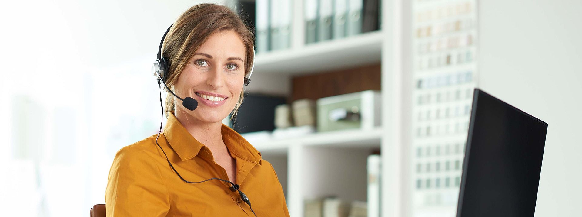 Representative image for customer service The picture shows a woman sitting at her computer and wearing headphones with an integrated microphone. It seems she's ready to take any customer request with ease.