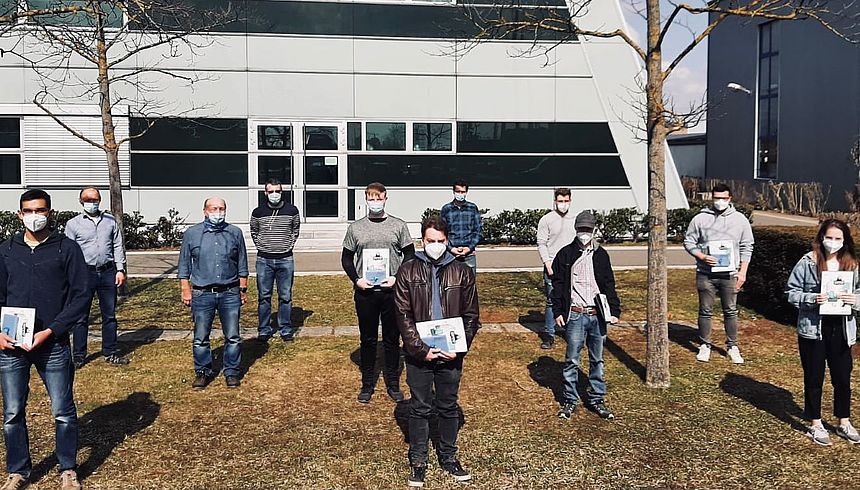 Gruppenbild der Absolventen Man sieht 12 Auszubildende und Ausbilder, welche Maske tragen und einen Sicherheitsabstand einhalten. Die Absolventen halten ihre Zeugnisse.
