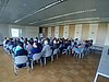 Eine sitzende Menge von etwa 30 Personen guckt auf ein vorne von einem Beamer angeworfenes Präsentationsbild