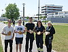 Fünf Auszubildende posieren mit ihren gefertigten Holzherzen. Sie stehen auf einem Weg vor einer Wiese und es ist sehr sonnig.