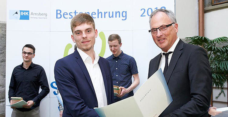 Man sieht zwei Männer in Anzügen auf einer Feier. Dem Auszubildenden bekommt Glückwünsche für seine Bestnoten.
