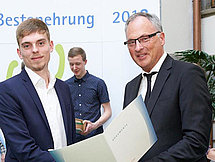 Man sieht zwei Männer in Anzügen auf einer Feier. Dem Auszubildenden bekommt Glückwünsche für seine Bestnoten.