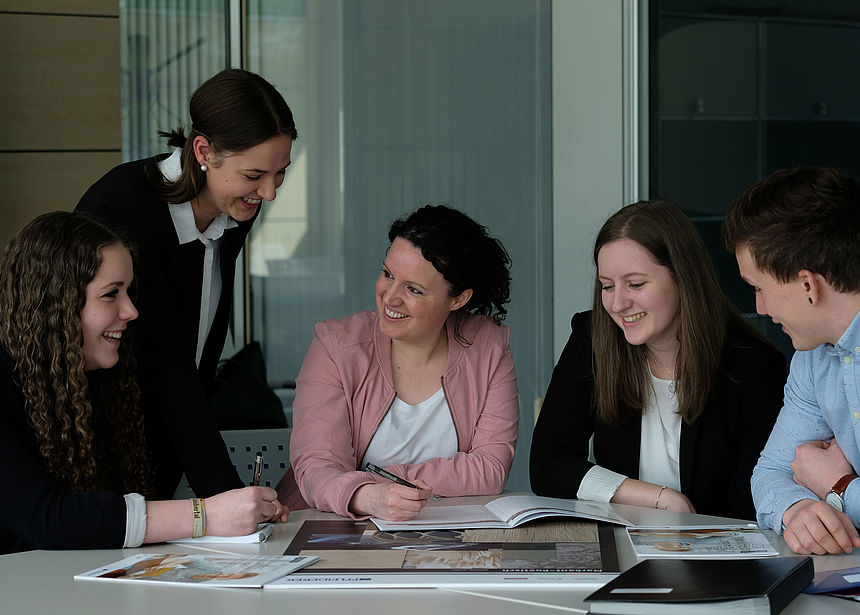 Mentoring The picture shows a group of young graduates and employees at a table together with a specialist from Pfleiderer. Some documents about Pfleiderer are lying in front of the persons.