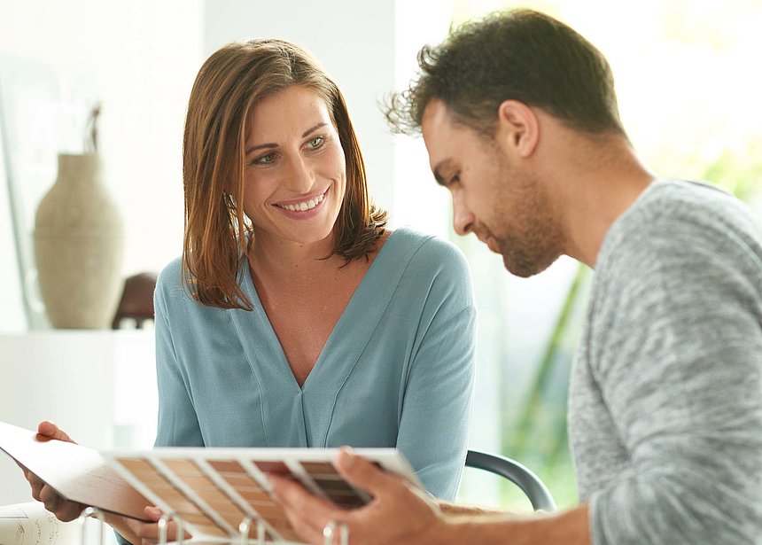 Personalentwicklung Das Bild zeigt eine Frau und einen Mann, die zusammen an einem Tisch sitzen. Vor ihnen liegen Mustermaterialien von Pfleiderer, von denen sie zu sprechen scheinen. Die Frau schaut den Mann direkt an und lacht, während er die Dokumente liest.