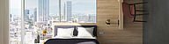 Das Bild zeigt ein Hotelzimmer mit einem Bett vor einem großen Eckfenster. Für Wände und Möbel werden verschiedene hochwertige Materialien verwendet.