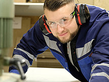 Das Bild zeigt einen Mann in Schutzkleidung, der große Maschinen bedient und direkt in die Kamera schaut