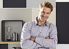 A man with blonde hair, dressed smart casually, is standing in a room with modern grey scale furniture and wooden accents. He's smiling into the camera. There's harsh lighting coming from the left.