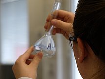Symbolbild Chemiekant Das Bild zeigt eine Frau, die einen Laborkittel und eine Schutzbrille trägt und einen Glaskolben in einem Labor betrachtet