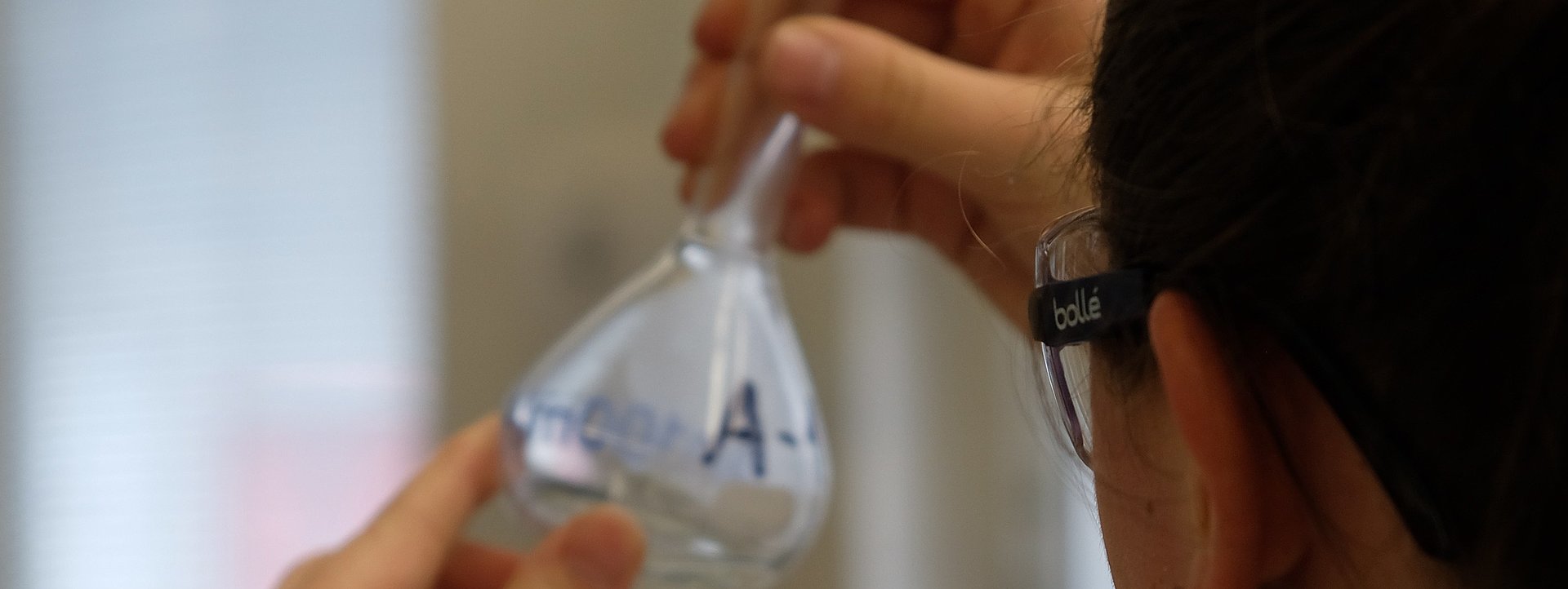 Symbolbild Chemiekant Das Bild zeigt eine Frau, die einen Laborkittel und eine Schutzbrille trägt und einen Glaskolben in einem Labor betrachtet